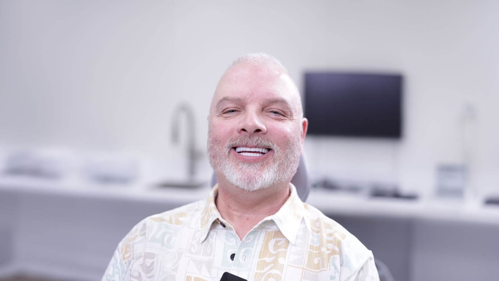 A smiling man with white hair and a beard sits in a brightly lit, modern office or clinic with blurred background equipment.
