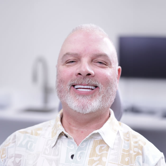 A middle-aged man with a trimmed white beard is smiling while seated indoors in a well-lit room with a blurred background.