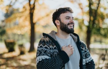 A cheerful man in a warm, patterned sweater stands amidst autumn trees, laughing freely. The sunlit background and fallen leaves capture the essence of an enjoyable, relaxed outdoor moment.