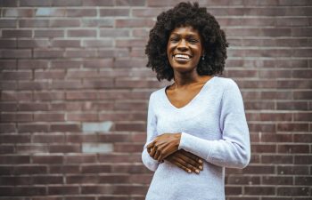 A cheerful woman with natural curly hair wearing a purple sweater, standing against a brick wall. The calming tones and vibrant smile create a warm and uplifting mood in an urban setting.